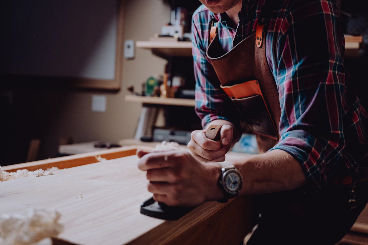 A woodworker in a leather apron hand-planing a timber board at a workbench